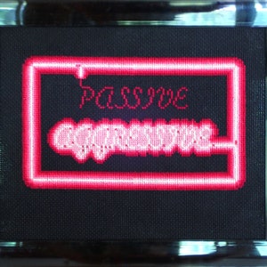 以下が含まれることがあります： 「PASSIVE AGGRESSIVE」とピンクのネオンサインが書かれた、額装されたクロスステッチの芸術作品です。