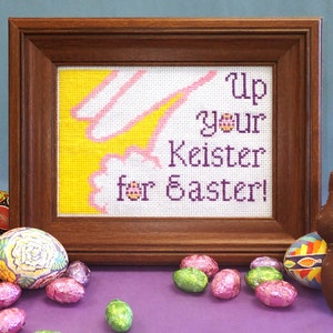 以下が含まれることがあります： ピンクのウサギと「Up your Keister for Easter!」というテキストが刺繍されたクロスステッチが、茶色の木製の額縁に入っています。額縁は、カラフルなイースターエッグとチョコレートのウサギで囲まれています。