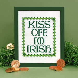 以下が含まれることがあります： 緑色の縁取りが施された白いクロスステッチ刺繍で、「Kiss Off I'm Irish」という文字が書かれています。刺繍は濃い緑色の額縁に収められ、緑色のシュレッダーペーパーと金貨で囲まれています。