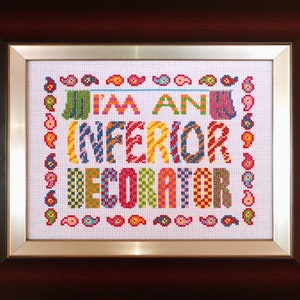 以下が含まれることがあります： 茶色のフレームに入ったクロスステッチのアートワークで、カラフルな文字で「I'M AN INFERIOR DECORATOR」と書かれています。 アートワークは、シルバーのボーダーとペイズリー柄で額装されています。