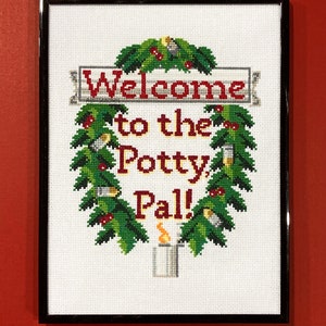 May include: A framed cross-stitch design with a green wreath and a sign that reads "Welcome to the Potty Pal!"