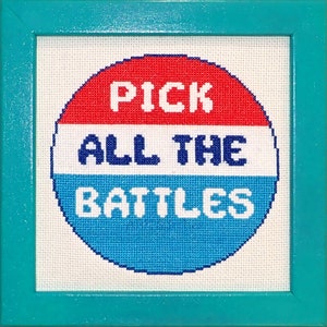 以下が含まれることがあります： 「Pick All The Battles」と書かれた赤、白、青の円が描かれた、額装されたクロスステッチのデザインです。
