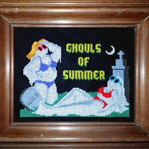 以下が含まれることがあります： 2人の幽霊が水着を着て「夏の悪霊」という文字と三日月が描かれた、額装されたクロスステッチのデザインです。