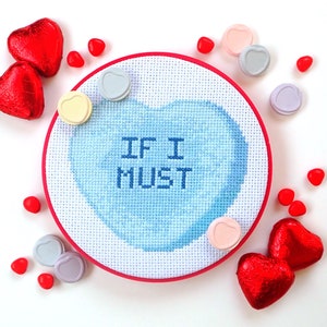 以下が含まれることがあります： 青いハートの中に「IF I MUST」という文字が書かれた、青と白のクロスステッチ刺繍のフープ。フープは赤とピンクのキャンディーハートで囲まれています。