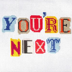 以下が含まれることがあります： 白い布地にクロスステッチで刺繍された「You're Next」というフレーズ。文字は赤、青、黄、緑など、さまざまな色で構成されています。