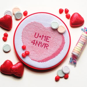 以下が含まれることがあります： ピンクと白のハート型の刺繍枠に、ピンクの文字で「U+ME 4EVER」と刺繍されています。枠の周りは赤と白のキャンディーハートで飾られています。