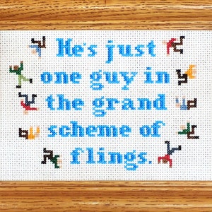 以下が含まれることがあります： 青い文字で「He's just one guy in the grand scheme of things.」と書かれた額装されたクロスステッチ。テキストは、小さくカラフルなピクセル化された人物で囲まれています。