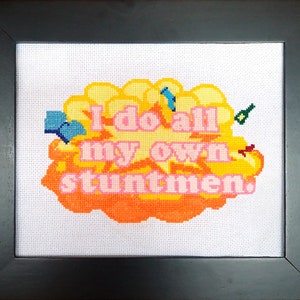 以下が含まれることがあります： 黄色とオレンジの雲の形をした額縁付きのクロスステッチで、ピンクの文字で「I do all my own stuntmen.」と書かれています。