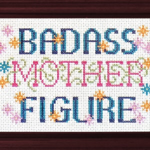 以下が含まれることがあります： 青い、ピンク、緑の文字で「BADASS MOTHER FIGURE」と書かれた、額縁に入ったクロスステッチ。文字の周りは、小さなカラフルな花で囲まれています。