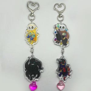 Op de afbeelding: Twee acryl sleutelhangers met cartoonkarakters. Eén sleutelhanger heeft een geel en blauw karakter, de andere een karakter met een penseel. Elke sleutelhanger heeft een hartvormige bedel en een zilverkleurige sluiting.