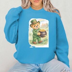 Könnte beinhalten: Ein hellblaues Sweatshirt mit einem St. Patrick's Day-Motiv. Die Grafik zeigt einen Teddybären in einem grünen Anzug und Hut, der einen Topf mit Gold hält. Ein Regenbogen und ein Haus sind im Hintergrund.