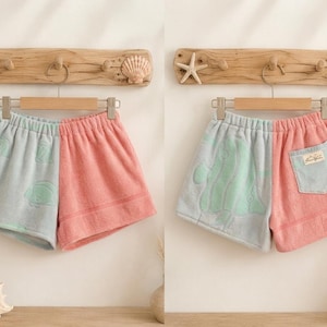May include: Two-toned terry cloth shorts featuring a split design with a light blue side and a coral pink side. The light blue side displays a subtle fish pattern, while the coral pink side includes a light blue patch pocket with a small label.
