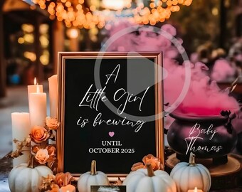 May include: A black framed sign with white text reading A Little Girl is brewing UNTIL OCTOBER 2025, featuring a small pink heart. Beside it sits a black cauldron with Baby Thomas written on it, white pumpkins, candles, and an ultrasound.