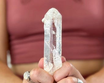May include: A clear quartz crystal with a textured surface is held between two hands. The crystal is elongated with a pointed tip. The background is a solid, muted red-pink colour.