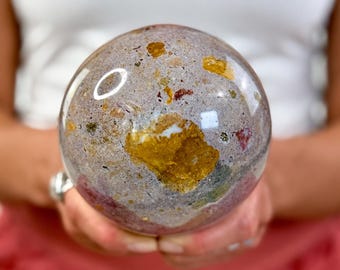 May include: A polished spherical stone featuring a mottled surface with a mix of lavender, grey, and white tones. The sphere is embedded with prominent, irregular patches of golden-yellow, deep red, and dark green mineral inclusions.