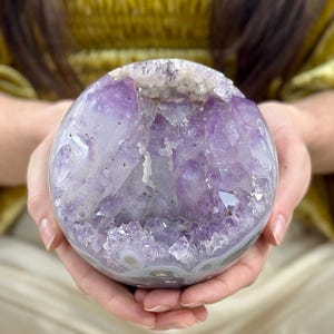 May include: A spherical amethyst geode, showcasing a vibrant purple crystal interior. The geode is held in open hands, revealing its polished exterior and natural crystalline formations. The geode's colors range from deep violet to pale lavender.