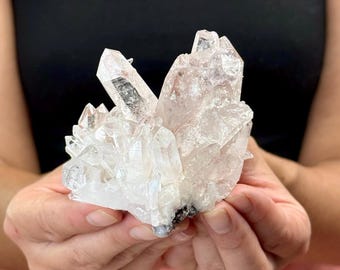 May include: A cluster of clear quartz crystals with a pale pink hue. The crystals are held in a person's hands, showcasing their natural, irregular shapes and varying sizes. The quartz has a translucent appearance.