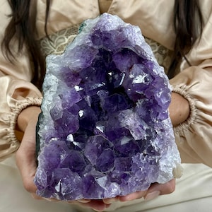May include: A large amethyst geode featuring deep purple, multifaceted crystal points clustered within a rough, light-colored rocky exterior. The crystalline structure displays varying shades of violet, ranging from pale lavender to dark, saturated purple hues.