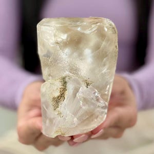 Pode incluir: Um grande espécime de cristal de quartzo amarelo-pálido, translúcido, com uma forma rugosa e irregular. A pedra apresenta fraturas internas, inclusões nubladas e uma superfície texturizada com manchas de depósitos minerais metálicos.