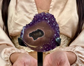 May include: A heart-shaped amethyst geode slice featuring deep purple crystalline edges surrounding a smooth, brownish-tan center with a dark, irregular cavity. The mineral specimen is mounted on a black metal stand for display purposes.