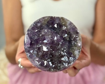May include: A spherical amethyst geode, showcasing a rich purple hue with crystalline formations. The geode's interior reveals sparkling crystal points and a natural, textured surface. The overall shape is round, and the geode is held in open hands.