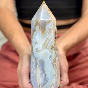 Pode incluir: Um obelisco de cristal polido e alto com uma ponta. O cristal apresenta uma mistura de cores azul claro, branco e castanho, com padrões e texturas naturais. O obelisco é segurado com as duas mãos.
