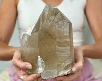 May include: A large, raw, smoky quartz crystal with a pointed top and a light brown color. The crystal is held in front of a person, showcasing its natural, textured surface and unique shape. A beautiful example of a natural crystal.