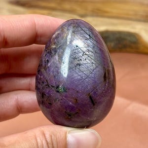Puede incluir: Una piedra preciosa pulida en forma de huevo, en tonos de morado intenso, con vetas negras y toques de verde. La piedra tiene una superficie lisa y reflectante, y se sostiene en una mano. El objeto es decorativo.