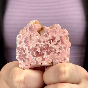 Könnte beinhalten: Ein Querschnitt eines rosa und weißen Rhodochrosit-Edelsteins. Der Stein hat ein geschichtetes Aussehen mit einer Mischung aus hellrosa und weißen Mustern. Die rauen Kanten des Steins sind hellbraun.