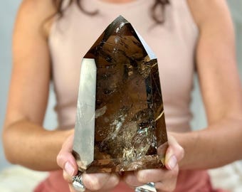 May include: A large, faceted smoky quartz crystal point. The crystal is a deep brown color with internal inclusions and a polished surface. The crystal is held in front of a person.