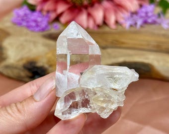 Puede incluir: Un grupo de cristales de cuarzo transparente, con un cristal grande y puntiagudo y cristales más pequeños adheridos. El cuarzo tiene una apariencia translúcida con sutiles inclusiones internas. El fondo presenta flores rosas y moradas.
