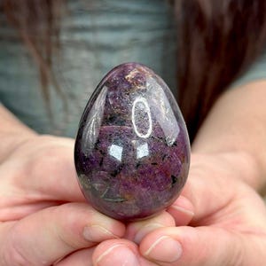 Puede incluir: Una gema pulida en forma de huevo, de un tono púrpura intenso, salpicada de inclusiones más oscuras. La piedra se sostiene en manos abiertas, mostrando su superficie lisa y reflectante. La forma y el color sugieren un objeto decorativo o un cristal curativo.
