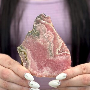 Könnte beinhalten: Ein polierter, roher Rhodochrosit-Stein mit geschichtetem Aussehen. Der Stein weist Bänder in Rosa- und Weißtönen auf, mit einigen Bereichen in Grün und Braun. Die Kanten sind rau und die Gesamtform unregelmäßig.