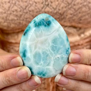 May include: A polished, teardrop-shaped gemstone with a striking pattern of white and turquoise blue. The stone is held in two hands, showcasing its smooth, glossy surface and unique color variations. The gemstone's natural beauty is highlighted by its intricate veining.