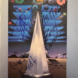 May include: A poster for The American Music Awards from January 27, 1992, with a geometric design and a silver obelisk. The poster has blue ink signatures and the Shrine Auditorium logo.