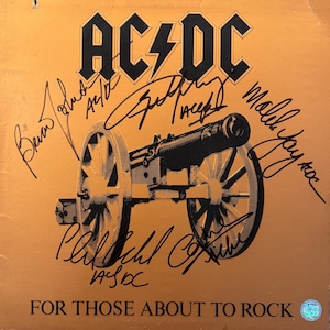 Puede incluir: Una portada de disco de vinilo cuadrada con fondo amarillo mostaza y una ilustración de un cañón negro. El texto AC/DC aparece en la parte superior y FOR THOSE ABOUT TO ROCK en la inferior. Varias firmas en tinta negra cubren la superficie.
