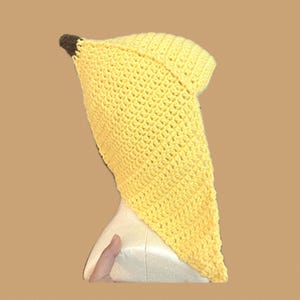 Puede incluir: Un sombrero amarillo tejido a ganchillo con forma de plátano, con una punta marrón. El sombrero está hecho de un material texturizado y tejido y está diseñado para ser usado en la cabeza. El sombrero es un artículo novedoso.