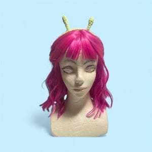 Puede incluir: Un busto de maniquí con una peluca rosa y una diadema verde claro con dos pequeñas decoraciones verticales en forma de antena. La peluca tiene flequillo y capas onduladas. El fondo es azul claro liso.