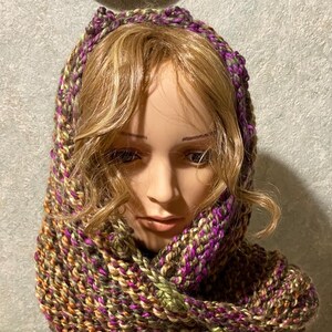 May include: A handmade, hooded scarf in a chunky knit design. The scarf features a blend of colors, including brown, green, orange, and purple. The hood is trimmed with a decorative edge. This cozy accessory is perfect for cold weather.