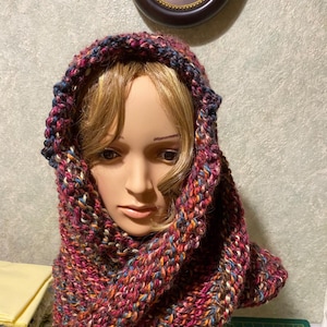 May include: A hooded scarf in a variety of colors, including red, blue, and orange, is draped over a mannequin head. The scarf is made of a thick, knitted yarn. The hood is pulled up over the head, and the scarf wraps around the neck.