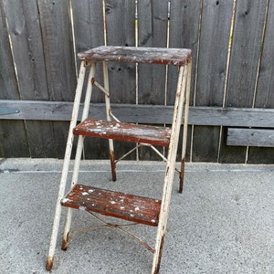 May include: A white metal step stool with three wooden steps. The steps are painted red and have white paint splatters on them. The stool is slightly rusted.