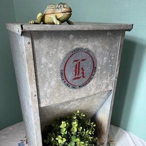 May include: A vintage metal feed dispenser with a red and white circular label that reads "Product of Circle K Kind, Inc. Mundelein, Ill. USA" and a large red "K" in the center. A small green frog figurine sits on top of the dispenser.