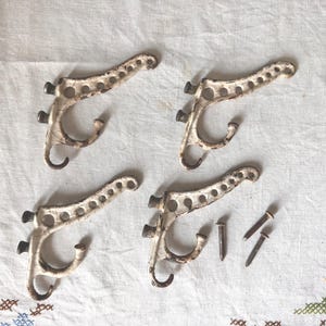 May include: Four antique white metal coat hooks with a distressed finish, arranged on a white cloth. Each hook has a curved design with a single hook and mounting hardware. Three nails are also present.