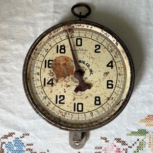 May include: A vintage, round, white dial hanging scale with black numbers and a black hand. The scale is made of metal and has a rusty patina. The scale is labeled "Capacity 20 lbs" and "Phila. MFG. CO. INC."