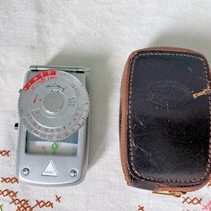 May include: A silver and black light meter with a circular dial and a leather case. The dial has markings for ASA, DIN, and shutter speeds. The case is brown and has a stitched edge.
