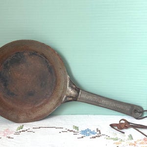 May include: An antique, round, cast iron griddle pan with a long handle. The pan has a dark, aged appearance with a brown interior and a silver handle. A leather strap is attached to the handle. The pan is likely used for cooking.