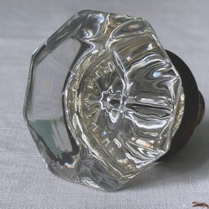May include: A clear, faceted glass door knob with a bronze-colored base. The knob has an octagonal shape and reflects light, creating a sparkling effect. This vintage-style hardware is suitable for doors and cabinets.