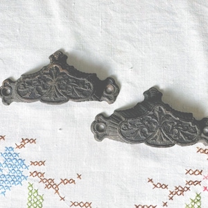 May include: Two antique, dark bronze drawer pulls with ornate floral designs. Each pull has a curved shape with decorative detailing and two holes for mounting. The pulls are set against a white background with embroidered floral patterns.