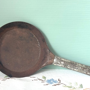 May include: An antique, round, cast iron griddle pan with a long handle. The pan has a dark brown, well-seasoned cooking surface and a silver-colored handle with embossed text. The handle has a hole for hanging.