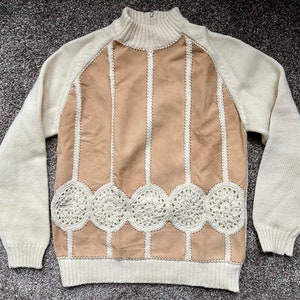 May include: A white sweater with a brown suede panel and crocheted trim. The suede panel has vertical stripes and a scalloped bottom edge.